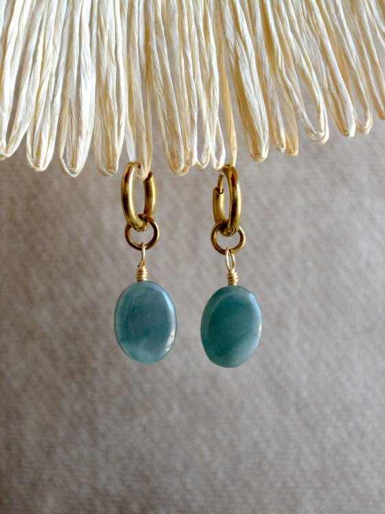 Aretes Belai Jade Princesa, argollas de acero dorado con dije de piedra jade ovalada, apoyados sobre lino junto a flores y hojas secas