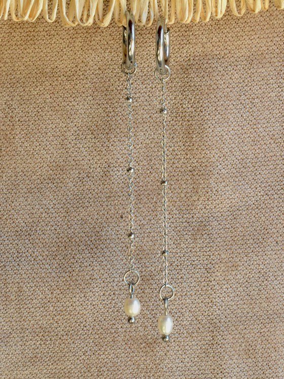 Aretes Belai Perla Colgante, argollas de acero plateado con cadena larga y fina terminada en una perla blanca colgante, apoyados sobre fibra de rafia y lino