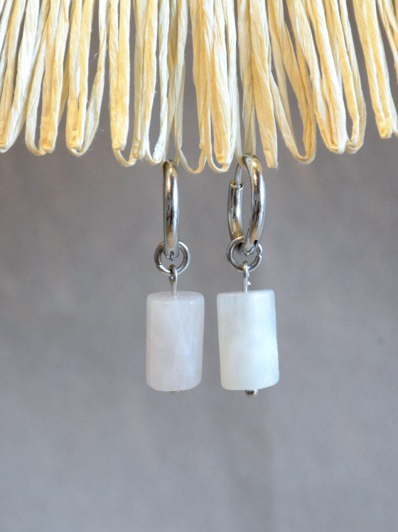 Aretes Belai Piedra Luna, argollas de acero plateado con dije colgante de piedra luna blanca en forma rectangular redondeada y pequeña bolita plateada en el extremo, apoyados sobre fibra de rafia y lino