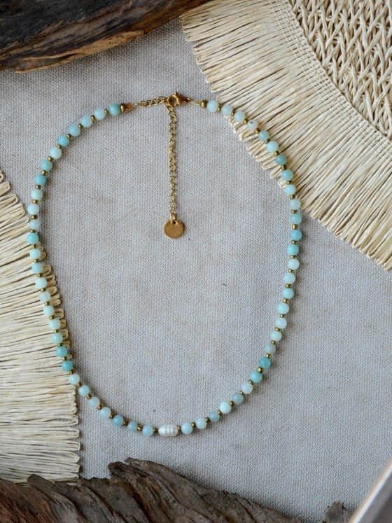 Collar Belai de cuentas redondas de amazonita celeste con pequeñas cuentas espaciadoras doradas, una perla blanca como acento asimétrico, broche de mosquetón dorado y cadena de extensión con dije redondo colgante, apoyado sobre lino junto a madera y fibra natural