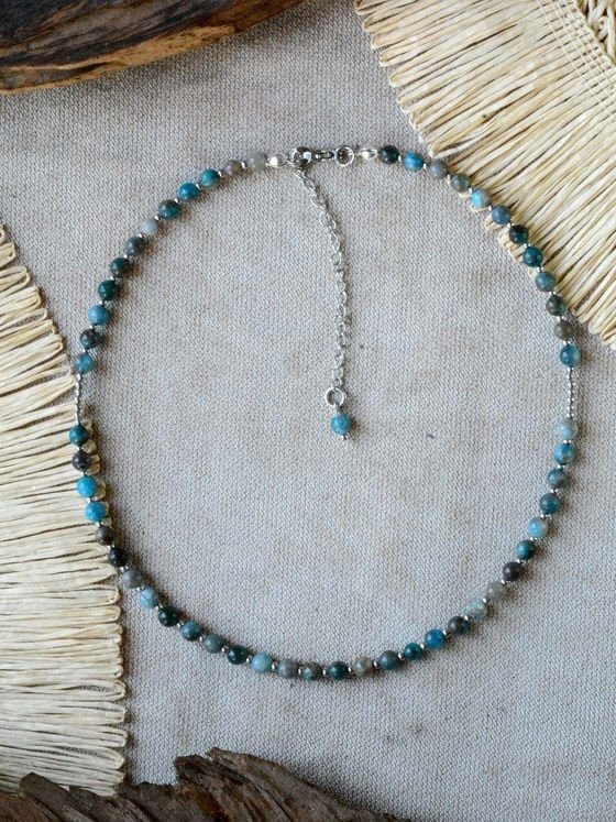 Collar Belai de cuentas pequeñas de apatita azul/verde y piedras labradorita gris, con broche de mosquetón plateado y cadena de extensión con dije de cuenta colgante, apoyado sobre lino junto a madera y fibra natural