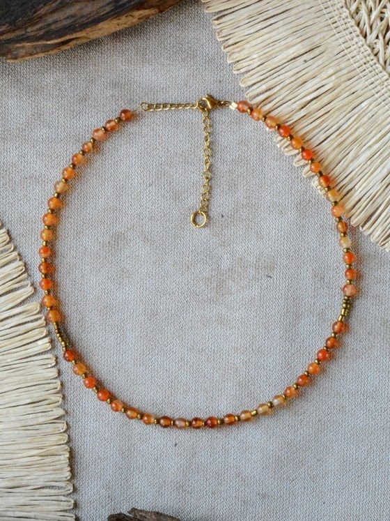 Collar Belai de cuentas de cornalina naranja con pequeñas cuentas espaciadoras doradas, broche de mosquetón dorado y cadena de extensión con dije redondo colgante, apoyado sobre lino junto a madera y fibra natural
