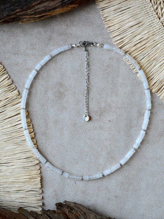Collar Belai de cuentas heishi de cuarzo blanco translúcido con pequeñas cuentas espaciadoras plateadas, broche de mosquetón plateado y cadena de extensión con dije redondo colgante, apoyado sobre lino junto a madera y fibra natural