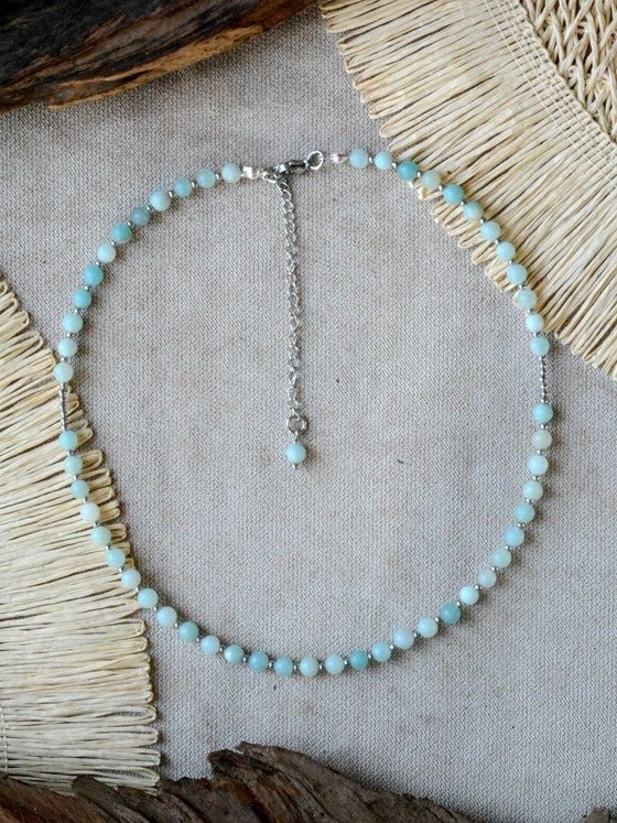 Collar Belai de Amazonita Plateada, cuentas redondas de amazonita celeste con pequeñas cuentas espaciadoras plateadas, broche de mosquetón plateado y cadena de extensión con una cuenta de amazonita colgante, apoyado sobre lino junto a madera y fibra natural