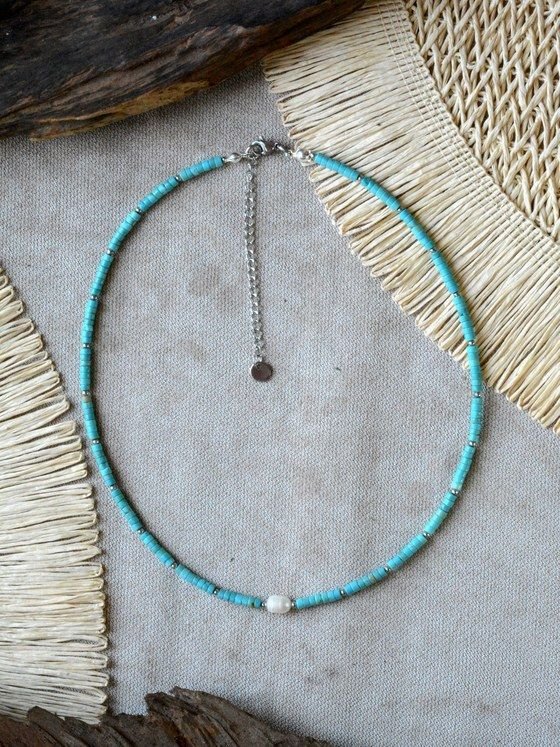 Collar Belai de cuentas heishi de turquesa con pequeñas cuentas espaciadoras plateadas, broche de mosquetón plateado, cadena de extensión con dije redondo colgante y una perla blanca como acento asimétrico, apoyado sobre lino junto a madera y fibra natural