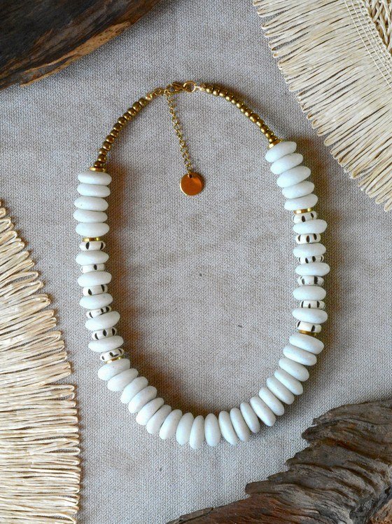 Collar Belai Medina Dorado, cuentas de vidrio reciclado color blanco crudo en forma de disco combinadas con cuentas de hueso decoradas con lunares negros, separadores y broche de mosquetón dorados, cadena de extensión con dije redondo colgante, apoyado sobre lino junto a madera y fibra natural