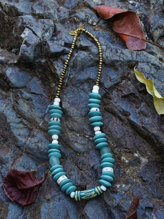 Collar Belai Esmeralda Krobo, cuentas de vidrio reciclado africano en tono verde esmeralda con detalles en bronce y hueso, apoyado sobre una piedra con hojas secas
