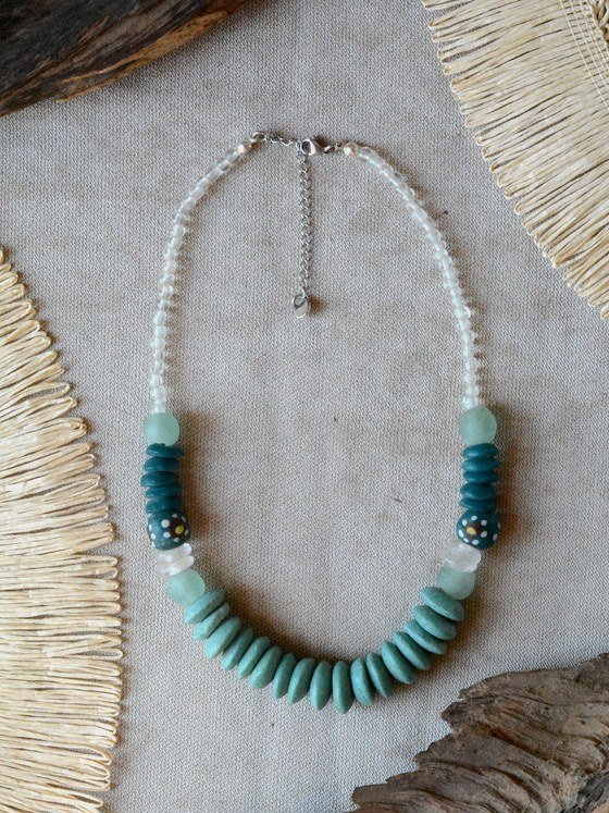 Collar Belai Esmeralda Plateada, discos de vidrio reciclado en tono verde esmeralda combinados con cuentas redondas pintadas a mano y cuentas de vidrio traslúcido, broche de mosquetón plateado y cadena de extensión con dije redondo colgante, apoyado sobre lino junto a madera y fibra natural