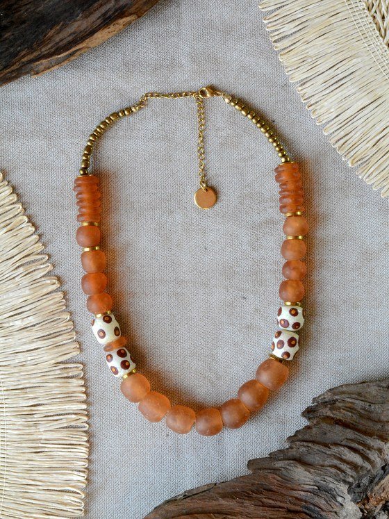 Collar Belai Sohori Dorado, cuentas de vidrio reciclado en tono ámbar con cuentas blancas de lunares y detalles en bronce, apoyado sobre una piedra con hojas secas