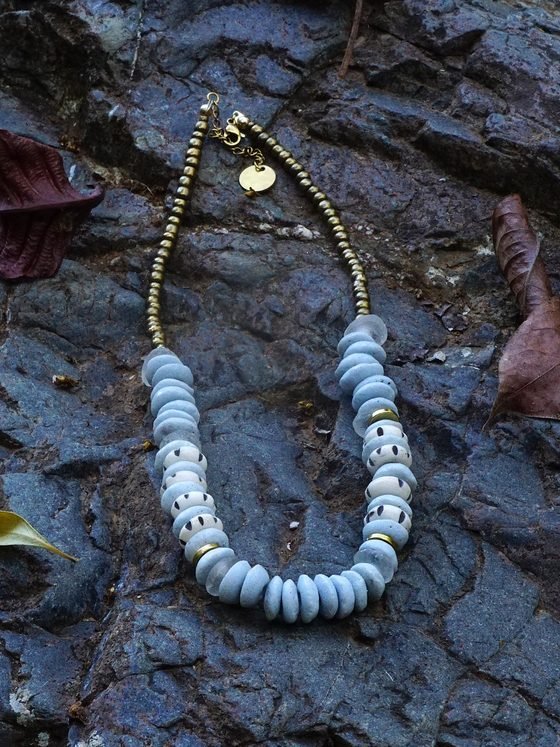 Collar Belai Tina Dorado, cuentas de vidrio reciclado en tonos celestes con detalles en bronce, apoyado sobre una piedra con hojas secas