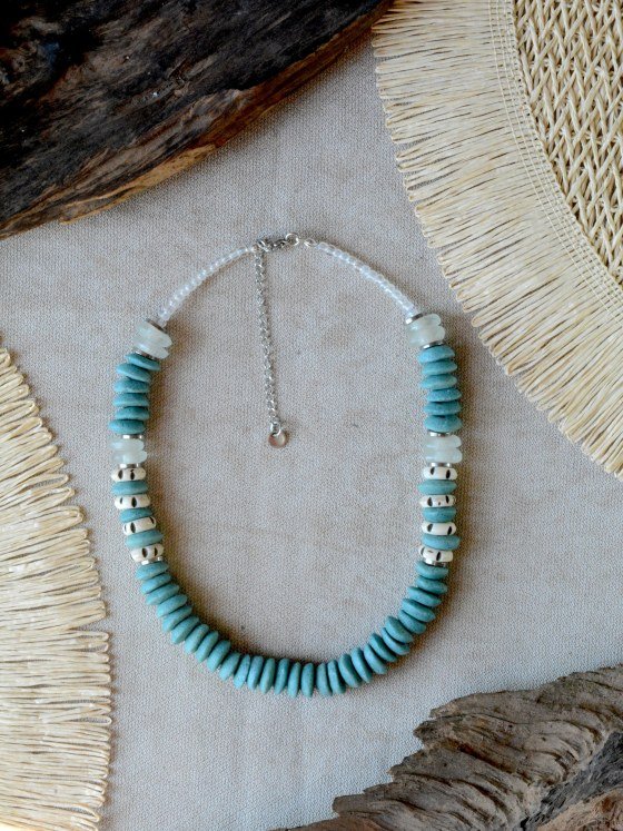 Collar Belai Tina Esmeralda, cuentas de vidrio reciclado en tono turquesa verde esmerilado combinadas con cuentas de hueso talladas color crudo con motivos pintados a mano en negro y vidrio traslúcido esmerilado, broche de mosquetón plateado y cadena de extensión con dije redondo colgante, apoyado sobre lino junto a madera y fibra natural