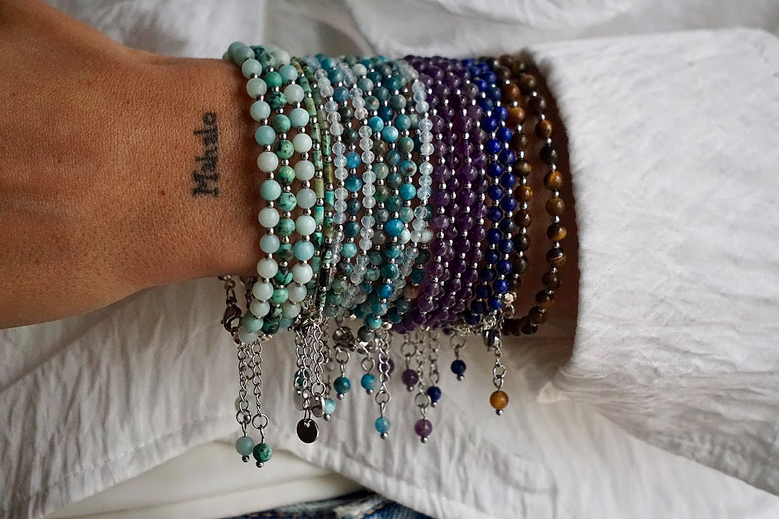 Antebrazo con muchas pulseras Belai de piedras naturales apiladas, en tonos turquesa, violeta, azul y marrón, sobre tela blanca