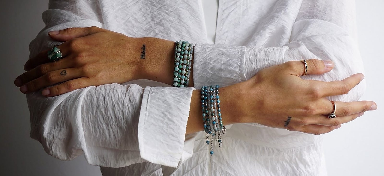 Manos cruzadas luciendo varias pulseras Belai de piedras naturales apiladas: amazonita y turquesa en un brazo, ónix, lapislázuli y perlas en el otro, sobre una camisa blanca de lino