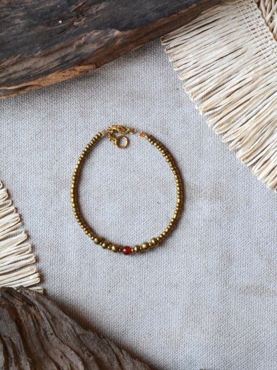 Pulsera Belai Vidrio y Cornalina, cuentas doradas de vidrio con una cornalina roja facetada, apoyada sobre lino junto a madera y fibra natural