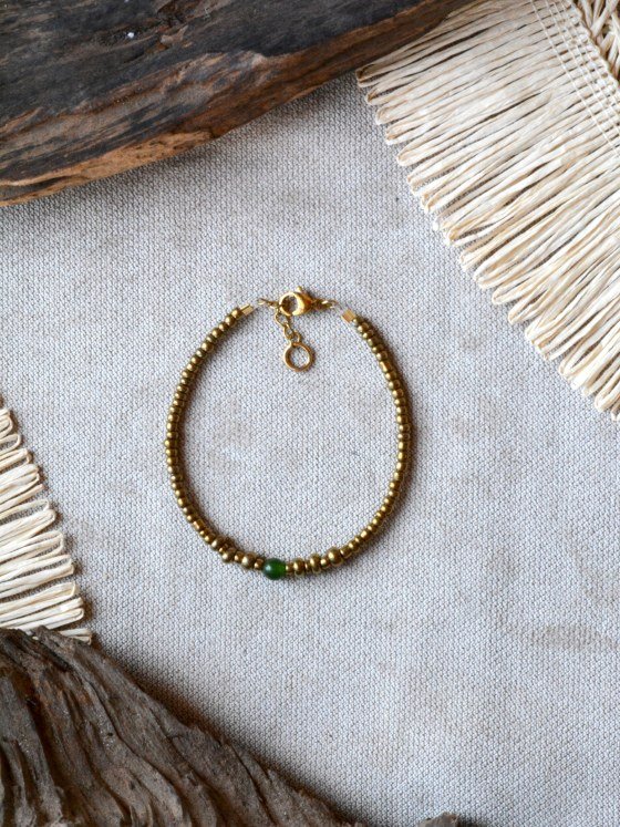 Pulsera Belai Vidrio y Jade Esmeralda, cuentas doradas de vidrio con una cuenta de jade verde esmeralda, apoyada sobre lino junto a madera y fibra natural
