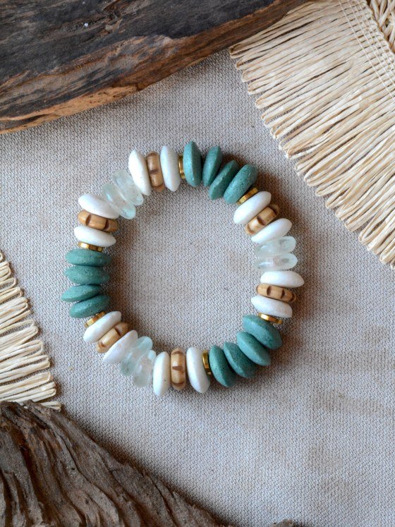 Pulsera Belai Esmeralda Dorada, cuentas en tono verde esmeralda y blanco con detalles en madera y anillos dorados, apoyada sobre lino junto a madera y fibra natural