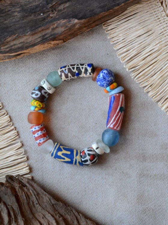 Pulsera Belai Krobo 2, cuentas de vidrio reciclado multicolor pintadas a mano al estilo Krobo, combinando vidrio traslúcido y cuentas pintadas en rojo, azul, marrón y amarillo, apoyada sobre lino junto a madera y fibra natural