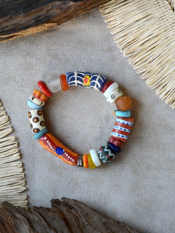 Pulsera Belai Krobo 4, cuentas de vidrio reciclado multicolor pintadas a mano con motivos variados: azul con telaraña y punto amarillo/rojo, blanco con ojos y lunares naranja, blanco con círculos marrón y naranja, naranja y rojo con líneas blancas, rojo/blanco a rayas con lunares azules, verde con líneas onduladas blancas, vidrio traslúcido naranja y celeste, apoyada sobre lino junto a madera y fibra natural