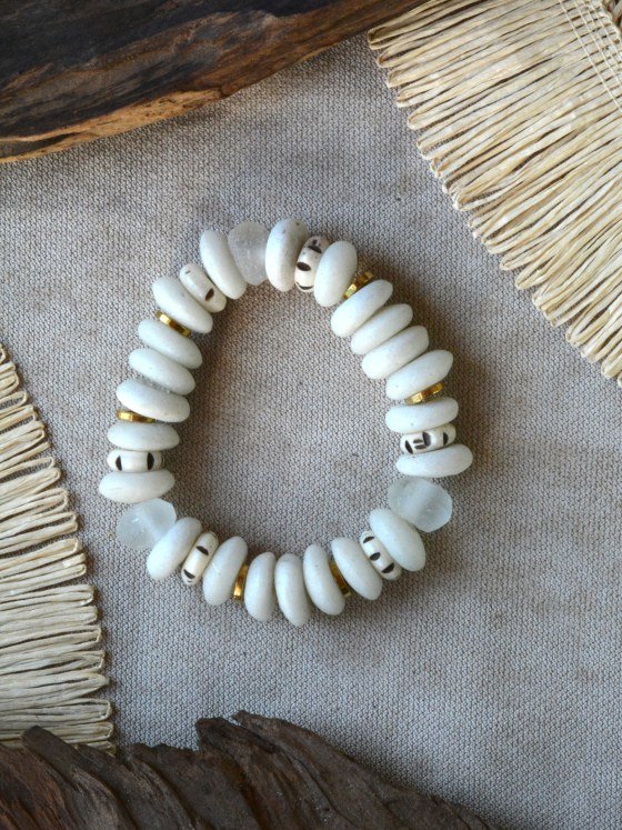 Pulsera Belai Medina, cuentas de hueso color blanco crudo combinadas con pequeñas cuentas talladas con motivos pintados a mano, vidrio traslúcido y anillos dorados, apoyada sobre lino junto a madera y fibra natural