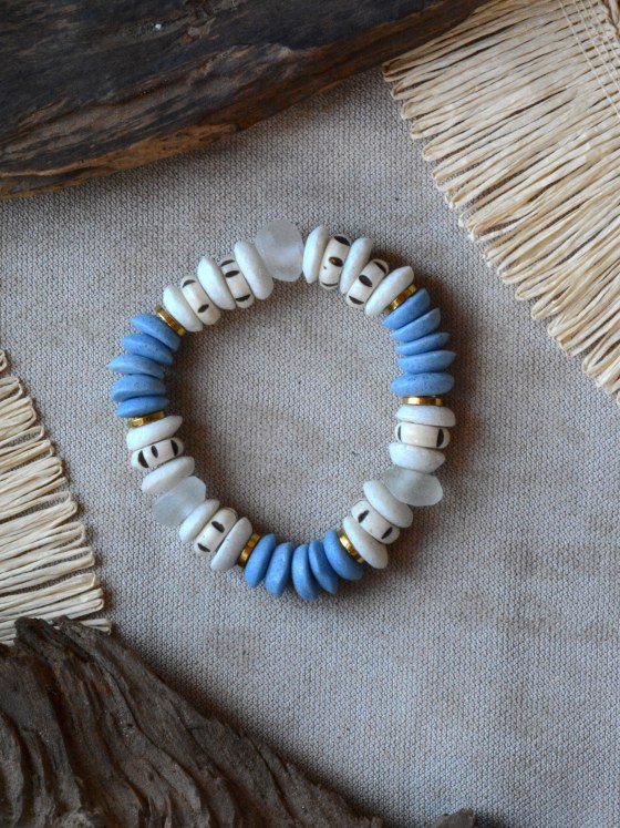 Pulsera Belai Tina Azul, cuentas de hueso color crudo con motivos pintados a mano combinadas con cuentas de vidrio reciclado azul, vidrio traslúcido y anillos dorados, apoyada sobre lino junto a madera y fibra natural
