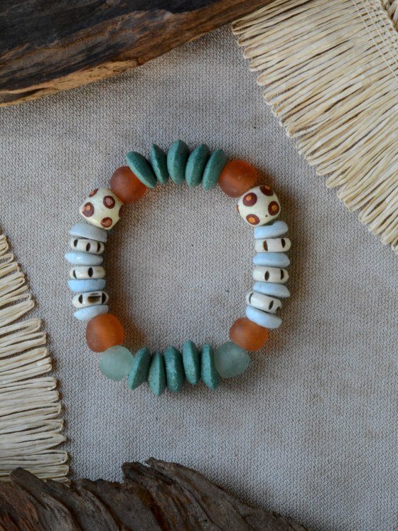 Pulsera Belai Tina Sohori, cuentas Krobo redondas color crudo con motivos tipo ojo pintados a mano en tonos marrón y naranja combinadas con cuentas de hueso talladas y cuentas celeste pálido, vidrio reciclado esmerilado naranja y traslúcido verde-mar, apoyada sobre lino junto a madera y fibra natural