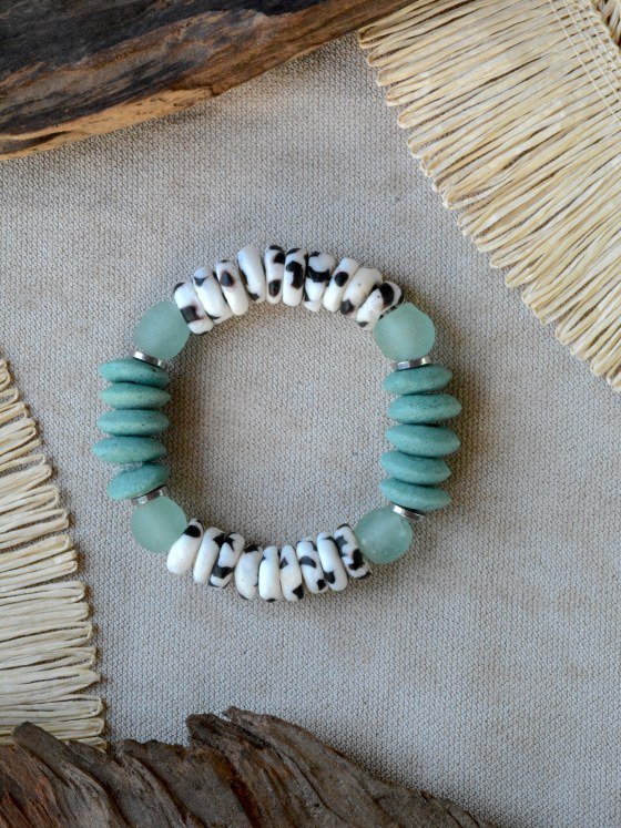 Pulsera Belai Zebra Esmeralda, cuentas blancas con motivos negros estilo animal print combinadas con cuentas de vidrio reciclado verde esmeralda, vidrio traslúcido celeste y anillos plateados, apoyada sobre lino junto a madera y fibra natural